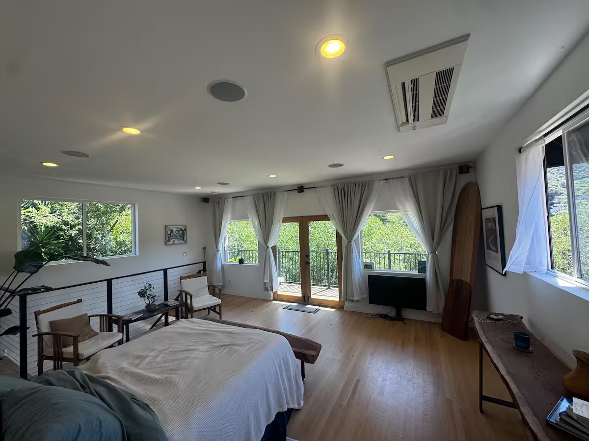 Wide view of the upstairs master bedroom with tall windows, sheer curtains, and reading chairs by the balcony doors